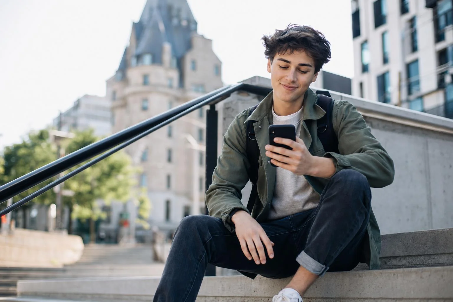 A casual young man sitting on outdoor city steps, glancing at his phone, relaxed and approachable.