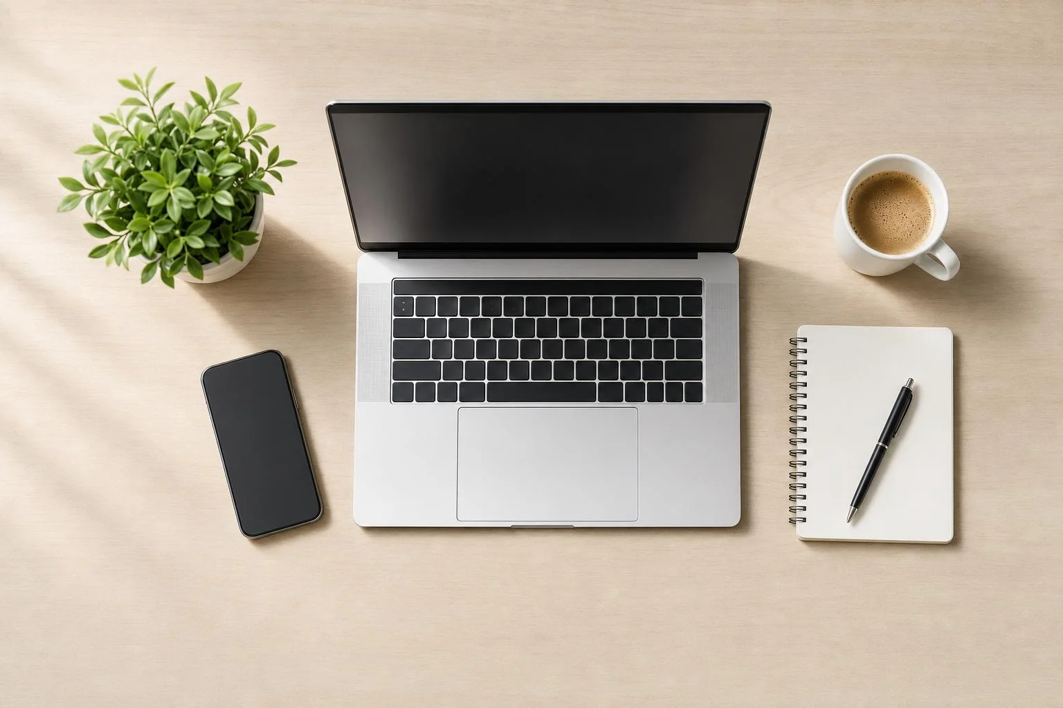 Bureau vu de haut sur bois clair : ordinateur portable, téléphone, plante, café et carnet, dans une lumière vive et aérée.