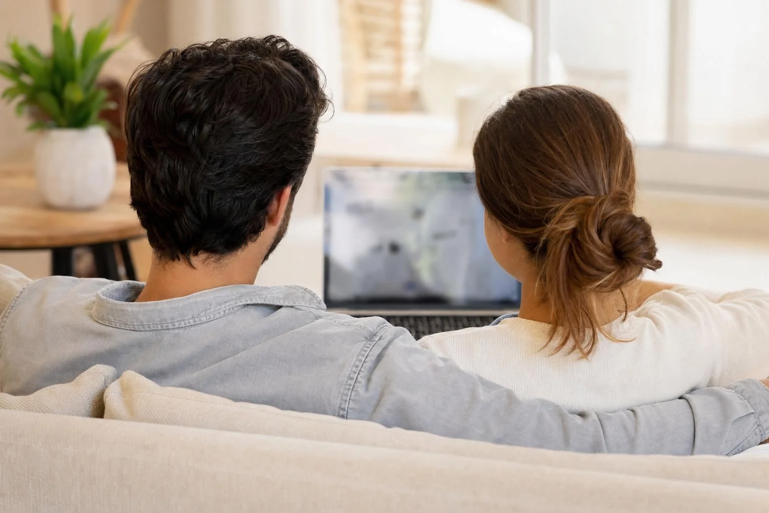 Un jeune couple détendu, vu de dos sur un sofa, qui regarde ensemble un ordinateur portable d'un air confiant.